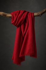 A rich ruby cashmere scarf with fringed edges draped over a rustic wooden branch against a dark gray studio background.